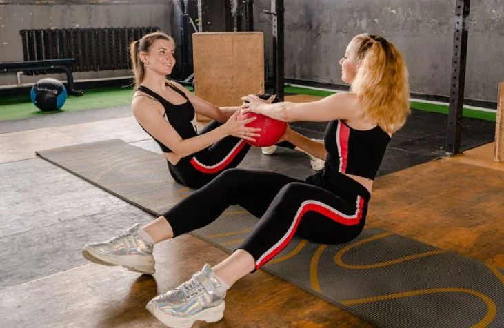 Two women performing a seated slam ball exercise together on workout mats.