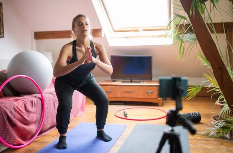 A woman in activewear performing a deep bodyweight air squat in a home living room, facing a camera on a tripod.
