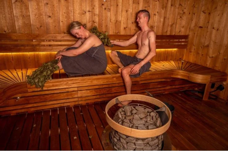 A man and a woman relaxing on wooden benches inside a traditional sauna holding birch twigs.
