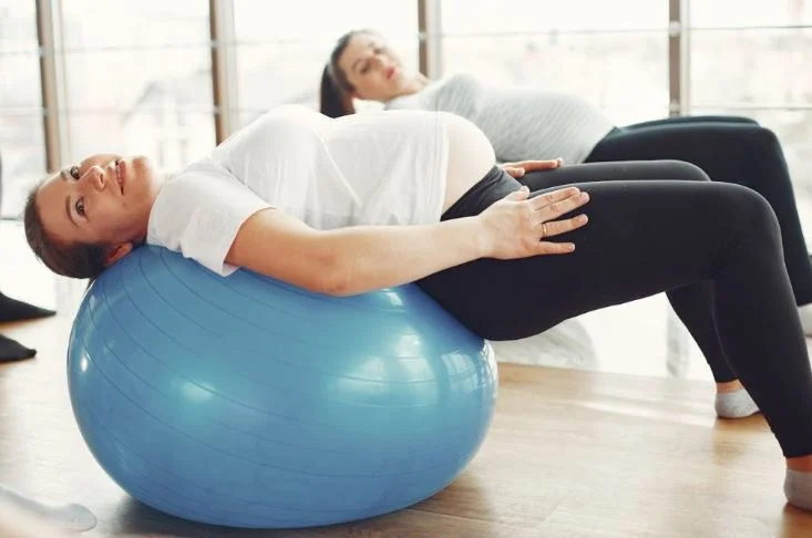 A pregnant woman lying comfortably on a yoga mat, leaning her back and shoulders against a large blue Pilates exercise ball while gently holding her abdomen.