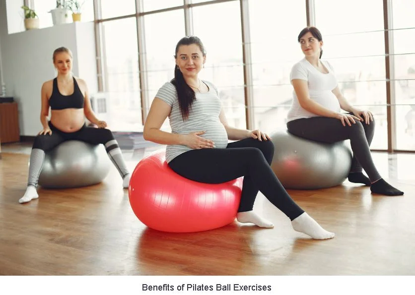 Three pregnant women sitting in a row on yoga mats in a bright, sunlit fitness studio, smiling and holding small Pilates balls behind their lower backs for support.